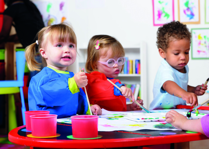 Group Of Toddlers Painting While Their Carer Tidies